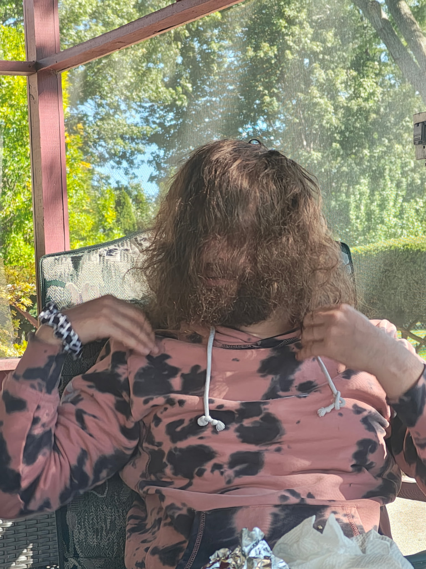David on a sunny screened porch, playfully hiding his face behind his long hair, wearing a pink tie-dye hoodie