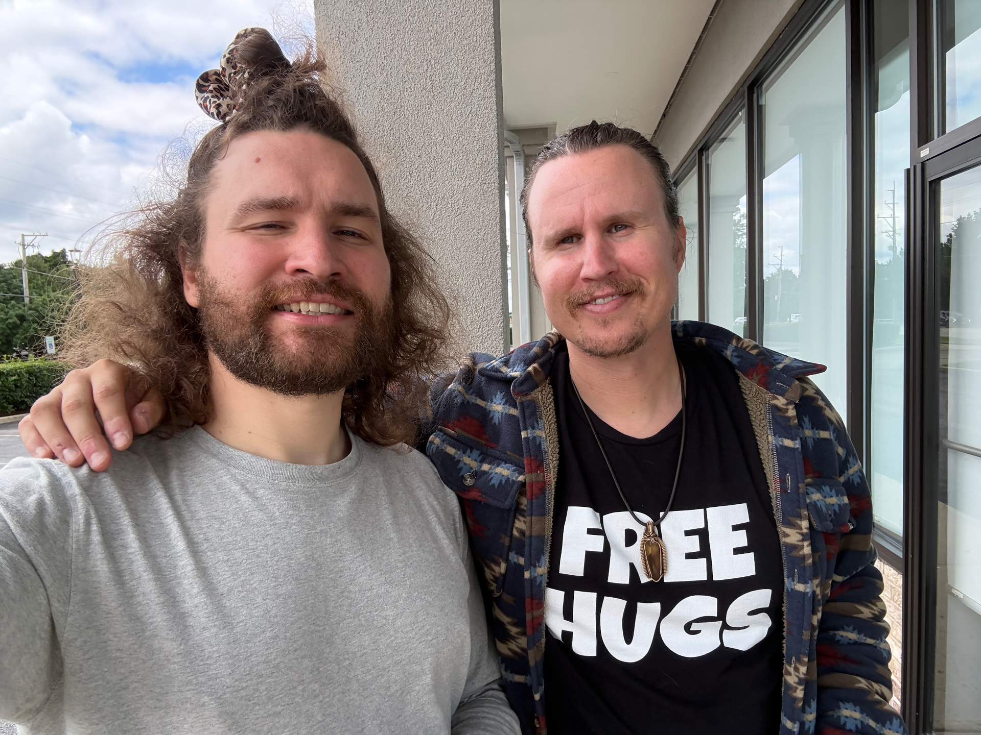 David and Kai smiling side by side outside a building, Kai in a Free Hugs shirt
