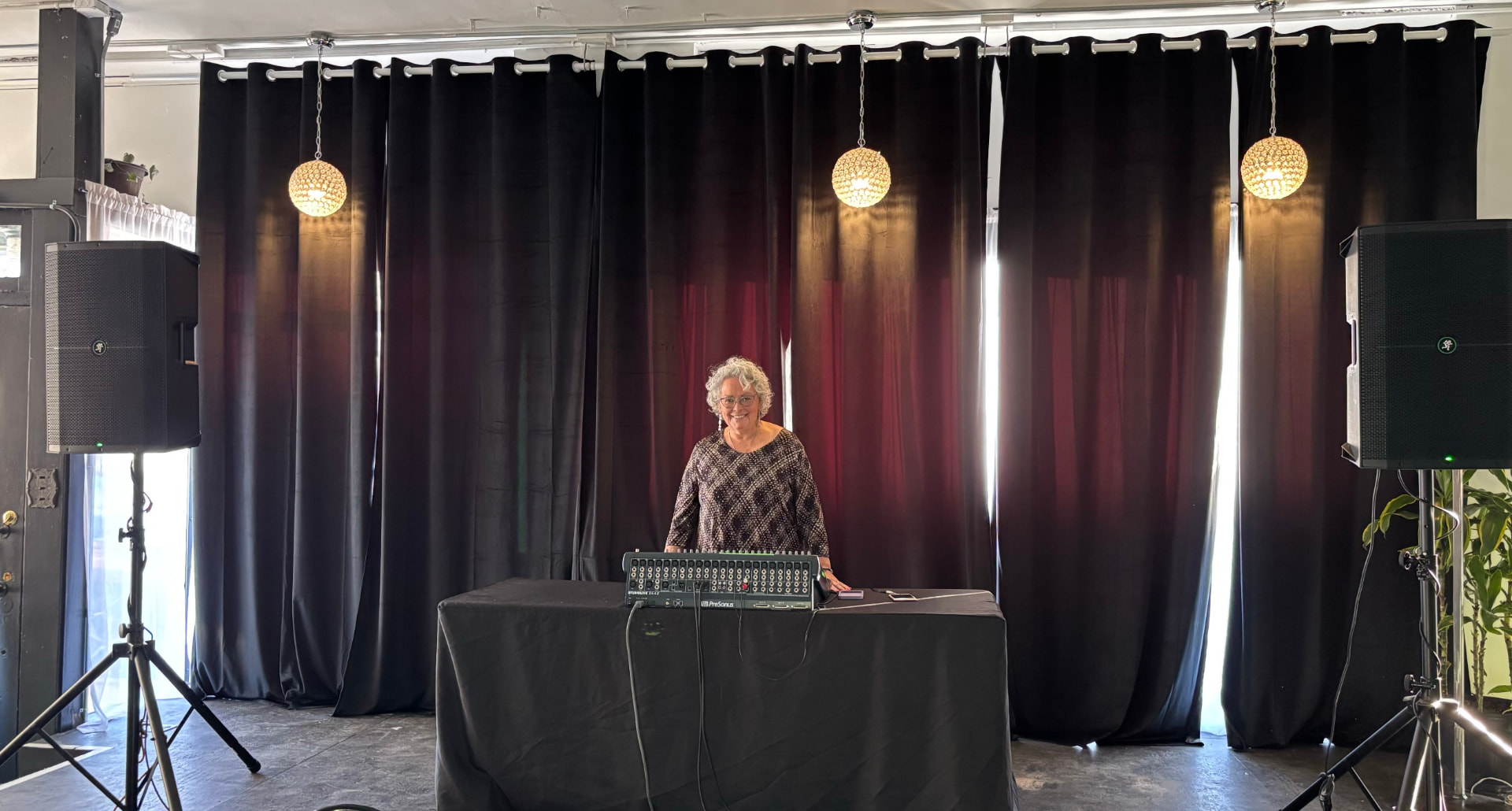 Yolanda Lozano standing behind a mixing board on a black-draped table, flanked by speakers under three hanging globe lights